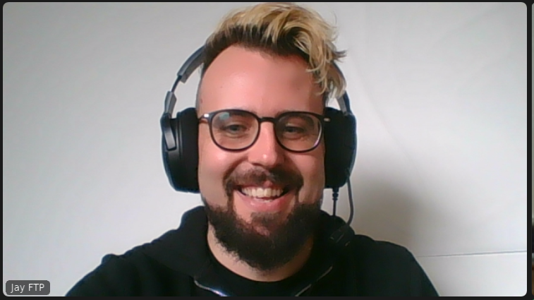 A person with a dark beard and hair with blonde highlights wearing glasses and a headset smiling.