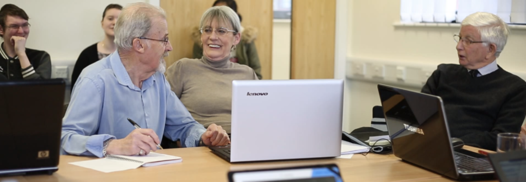 Older people sat at tables with laptop computers and mobile devices engaged in lighthearted conversation, with young volunteers in the background.