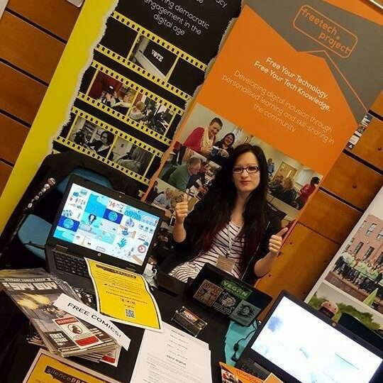 A woman with glasses and long dark hair sat at a promotional table with FreeTech Project information, while giving two thumbs up.