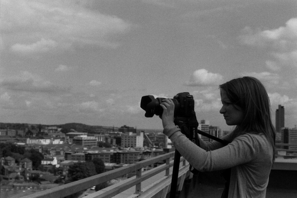 A woman with long dark hair looking into a video camera, composing a shot from a rooftop.
